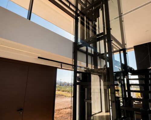 Γυάλινος ανελκυστήρας σε σύγχρονη luxury κατοικία με minimal μεταλλική σκάλα και μεγάλα κουφώματα αλουμινίου.